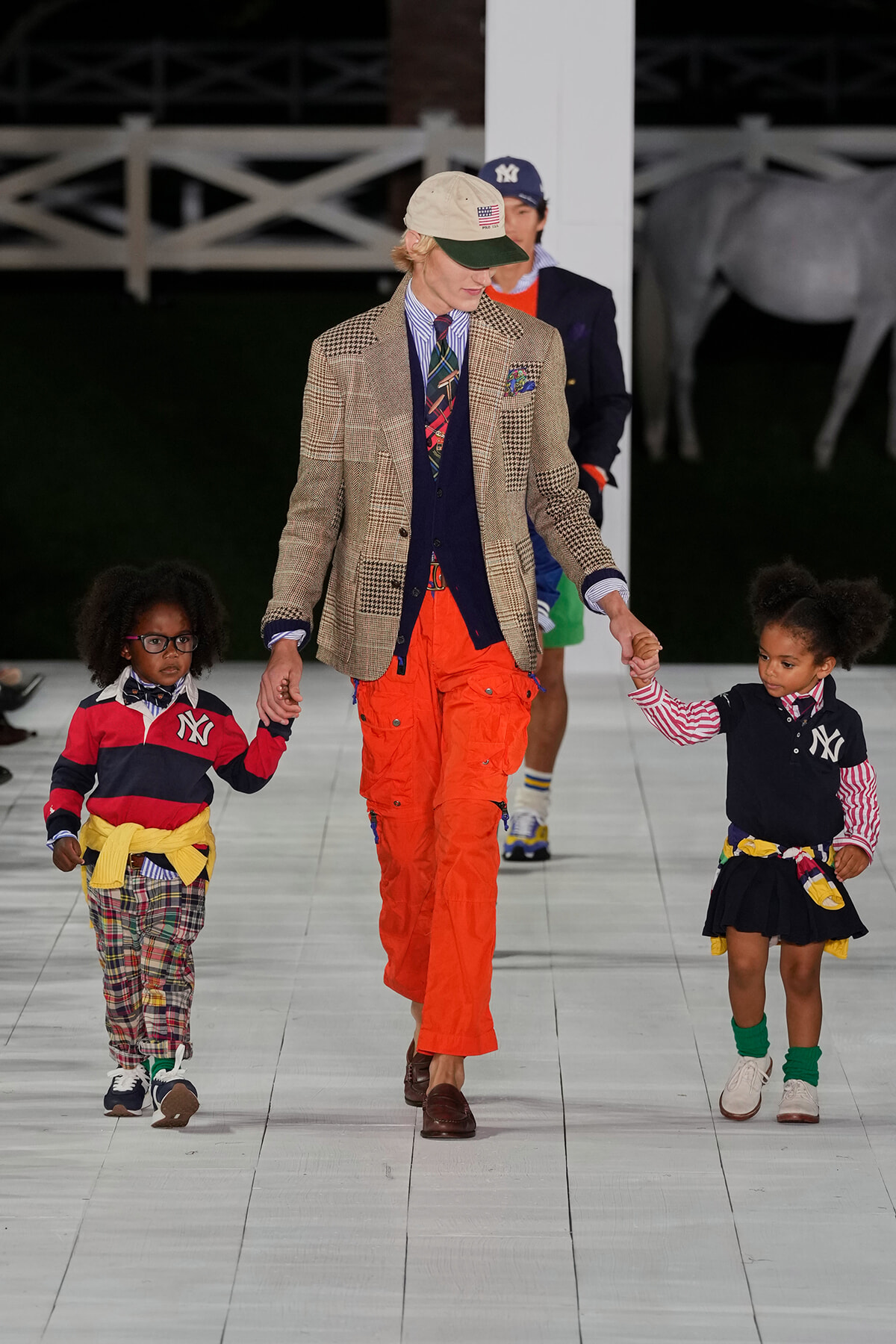 Adult model walks a white runway holding hands with two children in colorful outfits; beige plaid blazer, navy vest, striped shirt, and bright orange pants.