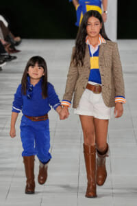Two young girls walk hand in hand down a fashion runway, wearing blue outfits and brown boots.
