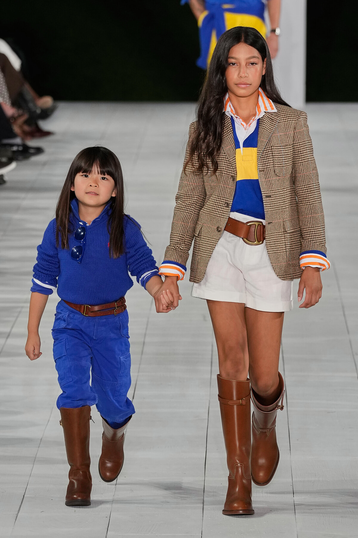 Two young girls walk hand in hand down a fashion runway, wearing blue outfits and brown boots.