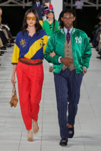 Two models walk a runway in bold streetwear: left is a woman in blue-yellow color-block polo, red high-waisted pants, tan sandals, and a small tan bag; right is a man in a green NY varsity jacket, plaid vest, pinstripe trousers, and loafers.