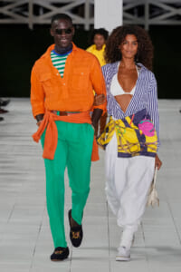 Two models walk a fashion show runway arm in arm: man in an orange shirt and green pants with a matching scarf, woman in a striped blue shirt over a white bikini top and white pants with a colorful wrap.