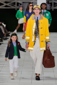 Adult and child model walk a fashion runway holding hands; the adult wears a bright yellow coat and tan cap, carrying a brown leather bag.