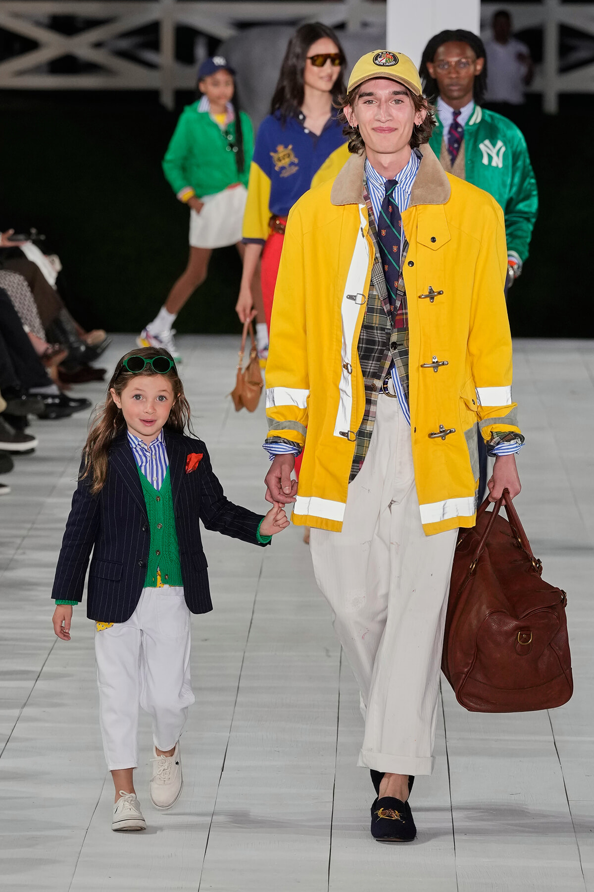 Adult and child model walk a fashion runway holding hands; the adult wears a bright yellow coat and tan cap, carrying a brown leather bag.