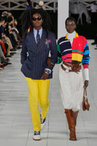 Two models walk the runway arm in arm: left, a man in a navy pinstripe double‑breasted blazer with a striped shirt, colorful tie, bright yellow trousers, and sneakers; right, a woman in a bold color‑blocked top, white turtleneck, white trousers, knee‑high brown boots, and a brown handbag.