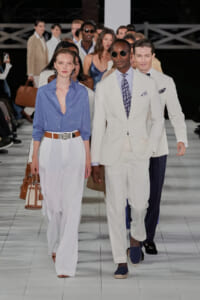 Models walk the runway in a fashion show; foreground features a woman in a light blue blouse with white wide-leg pants and a tan belt, alongside a man in a white suit with a patterned tie and sunglasses.