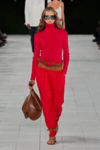 Model walking a runway in a red turtleneck and matching red pants, with a wide brown belt, a large brown bag, strappy sandals, and futuristic dark sunglasses.