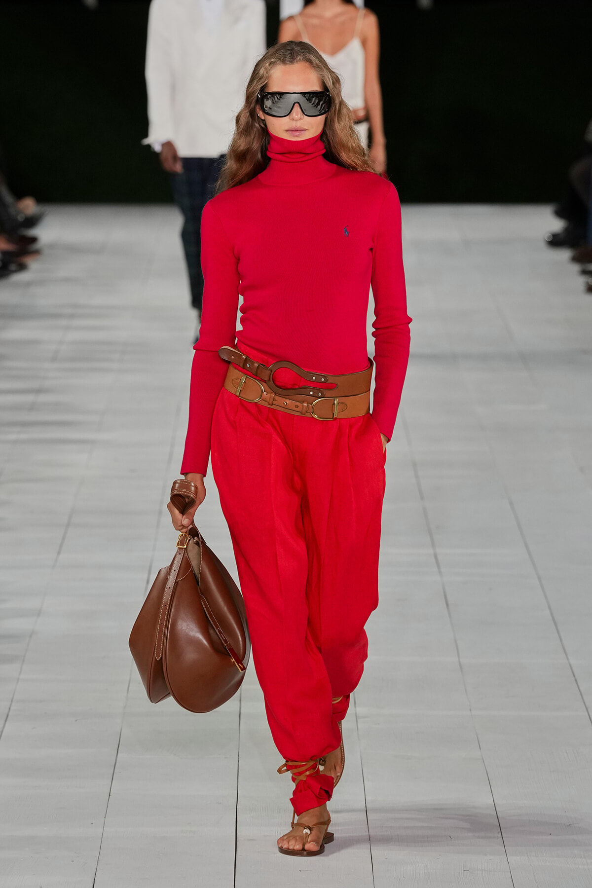 Model walking a runway in a red turtleneck and matching red pants, with a wide brown belt, a large brown bag, strappy sandals, and futuristic dark sunglasses.