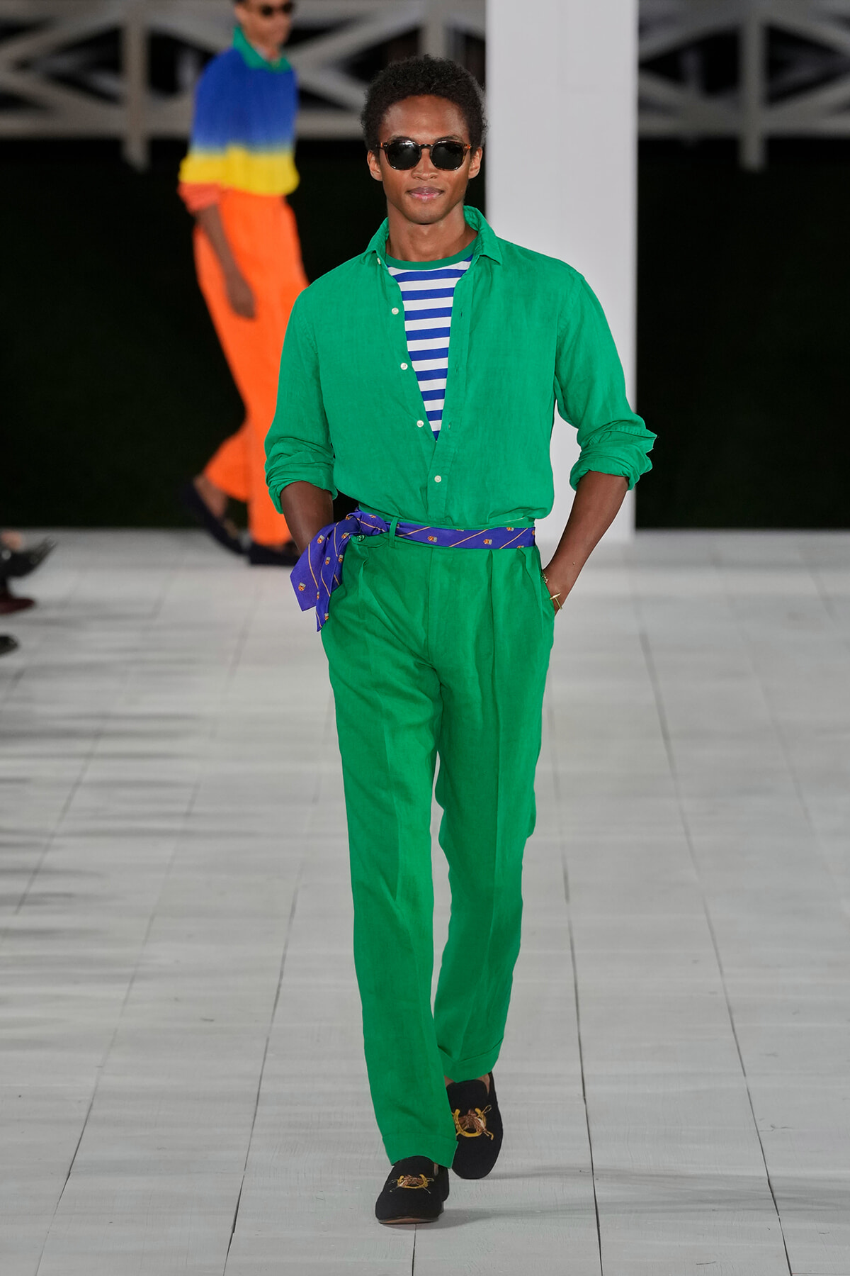 Male model walking a runway in a bright green linen suit, striped blue-and-white tee, and dark sunglasses.