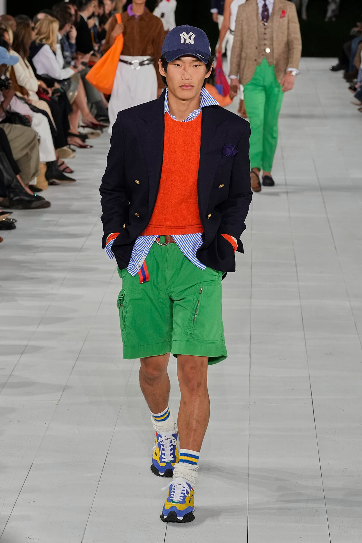 Male model walks a fashion show wearing a navy double-breasted blazer over an orange sweater and striped shirt, bright green cargo shorts, a New York Yankees cap, and colorful sneakers.
