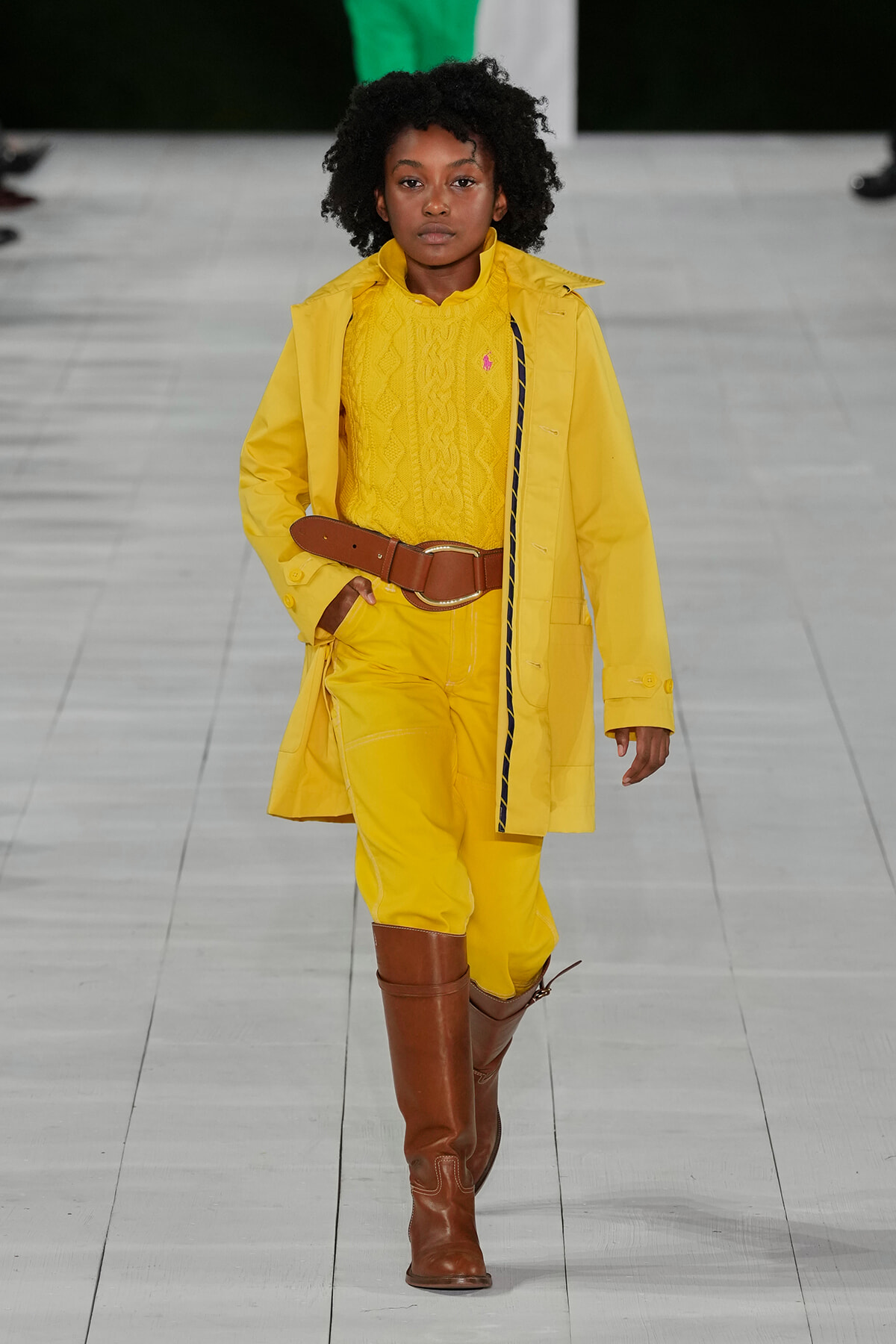 Model walks a fashion show in a bright yellow coat over a matching yellow sweater and pants, cinched with a wide brown belt and brown knee-high boots