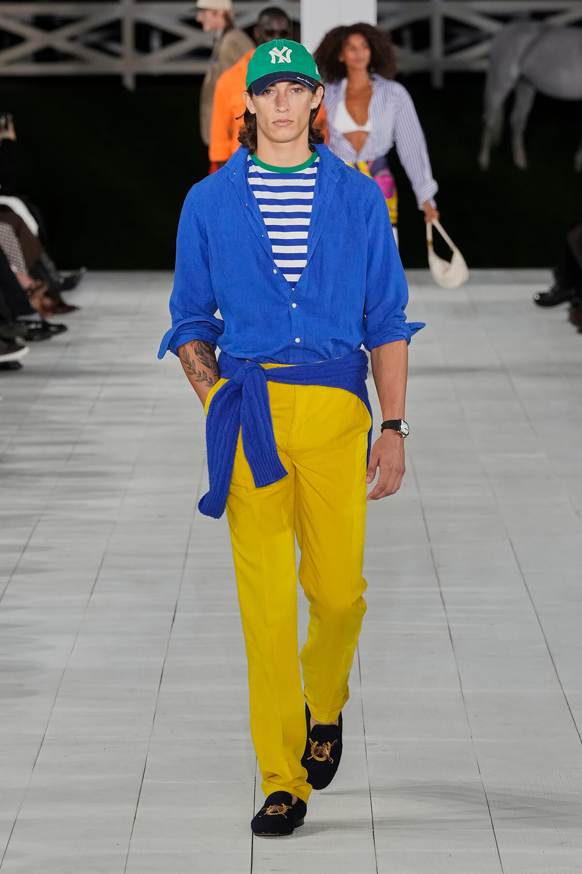 Male model walking runway in a blue button-down shirt over a striped tee, yellow pants with a blue sweater tied at the waist, green New York cap, and black loafers with gold embroidery.