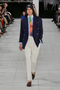 Male model walks a runway in a navy double-breasted blazer, striped shirt, colorful tie, white trousers, brown loafers, and a baseball cap with NY logo.