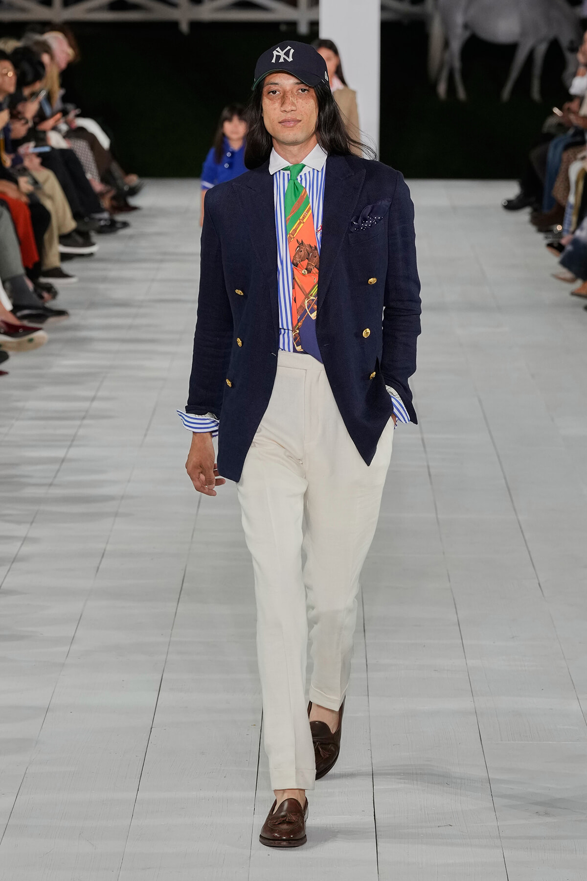 Male model walks a runway in a navy double-breasted blazer, striped shirt, colorful tie, white trousers, brown loafers, and a baseball cap with NY logo.
