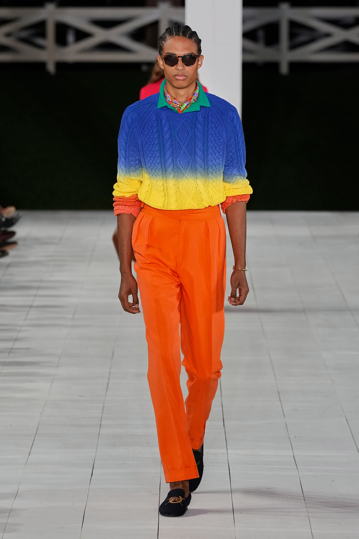 Model on runway wearing a blue–yellow–orange gradient knit sweater with a green polo collar, bright orange trousers, scarf, sunglasses, and loafers.