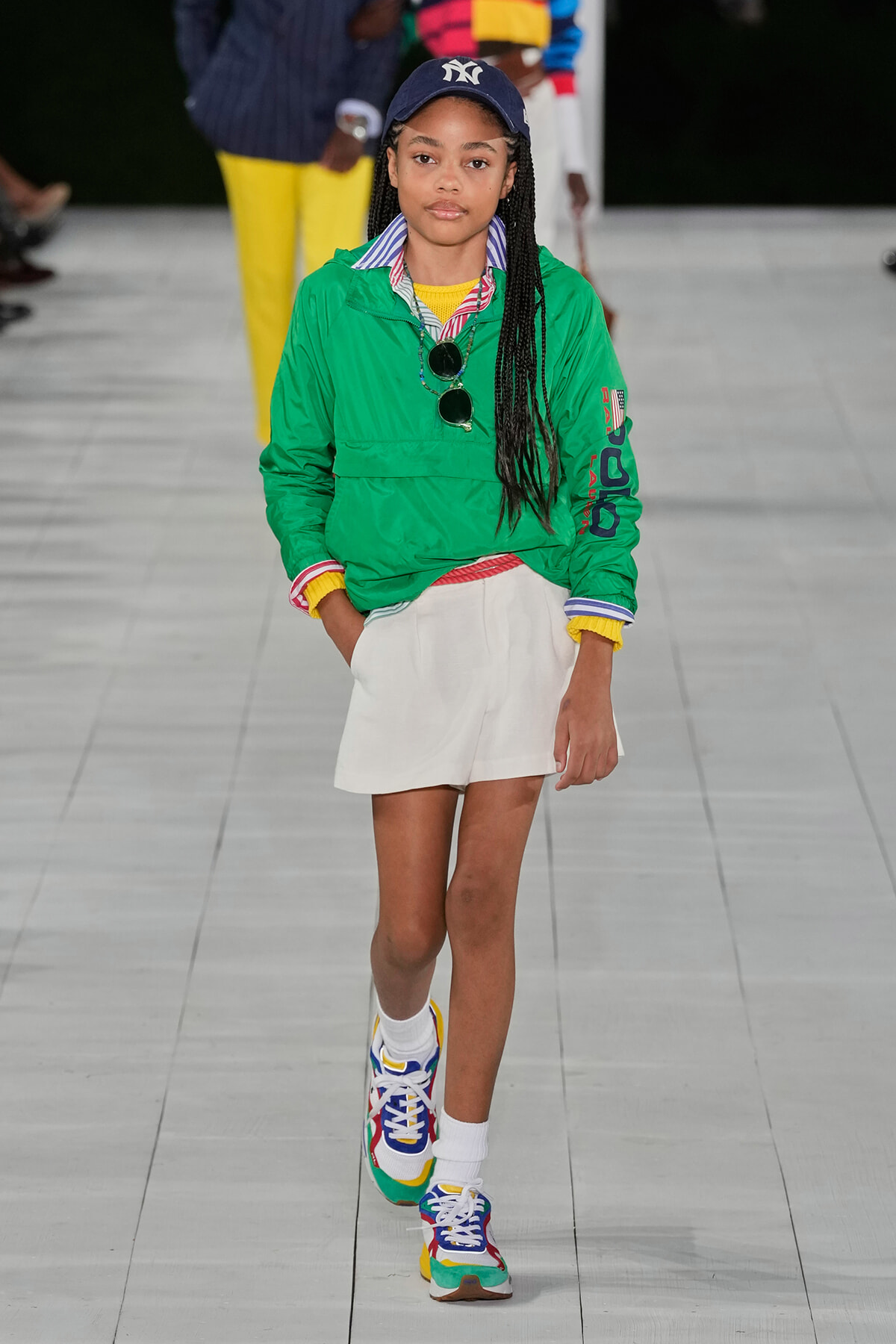 Model walking a runway in a bright green jacket, white shorts, colorful sneakers, and a navy Yankees cap.