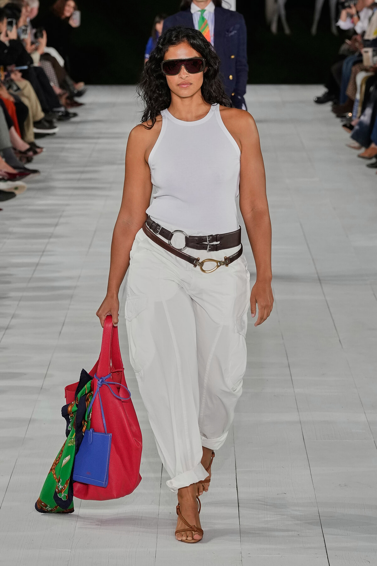Model on a runway wearing a white sleeveless top and white cargo pants, paired with two brown belts, brown sandals, oversized sunglasses, and a large colorful tote bag with a scarf accent.