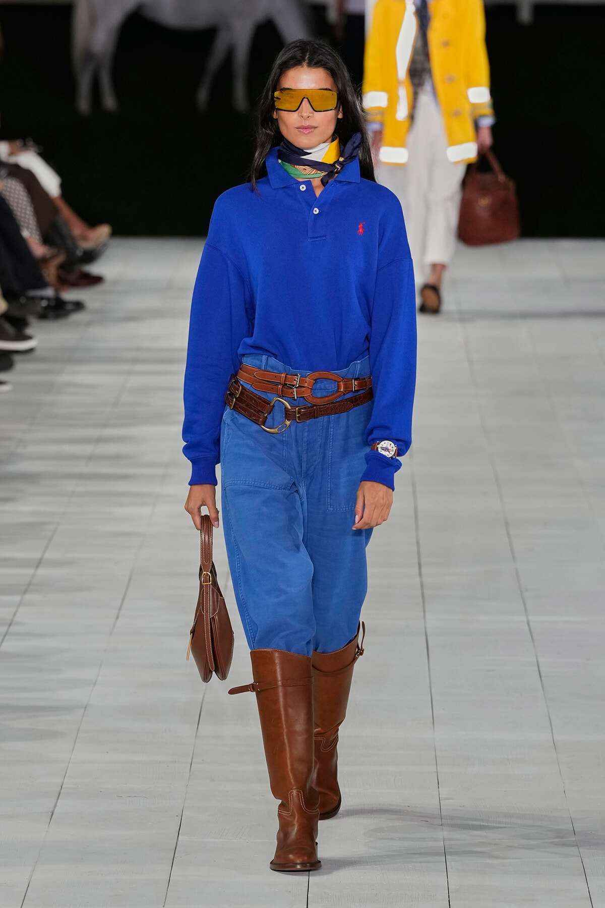 Model on a runway wearing a cobalt blue polo and matching blue pants, layered brown belts, tan knee-high boots, yellow visor sunglasses, scarf, and a brown handbag.