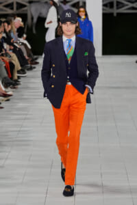 Male model on a runway wearing a navy double-breasted blazer with gold buttons, blue striped shirt, colorful scarf, bright orange trousers, navy loafers, and a New York Yankees cap.