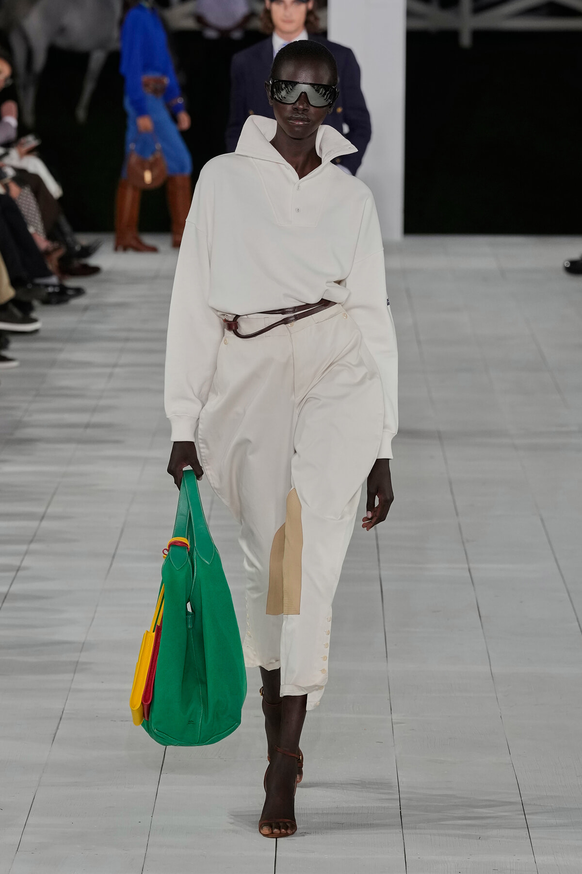 Runway model in an all-white oversized outfit with a brown belt, large sunglasses, and a bright green tote with yellow and red accents.