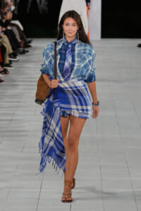 Model walking runway in blue plaid shirt paired with a matching blue plaid wrap skirt and tie, carrying a woven straw bag and wearing brown sandals.
