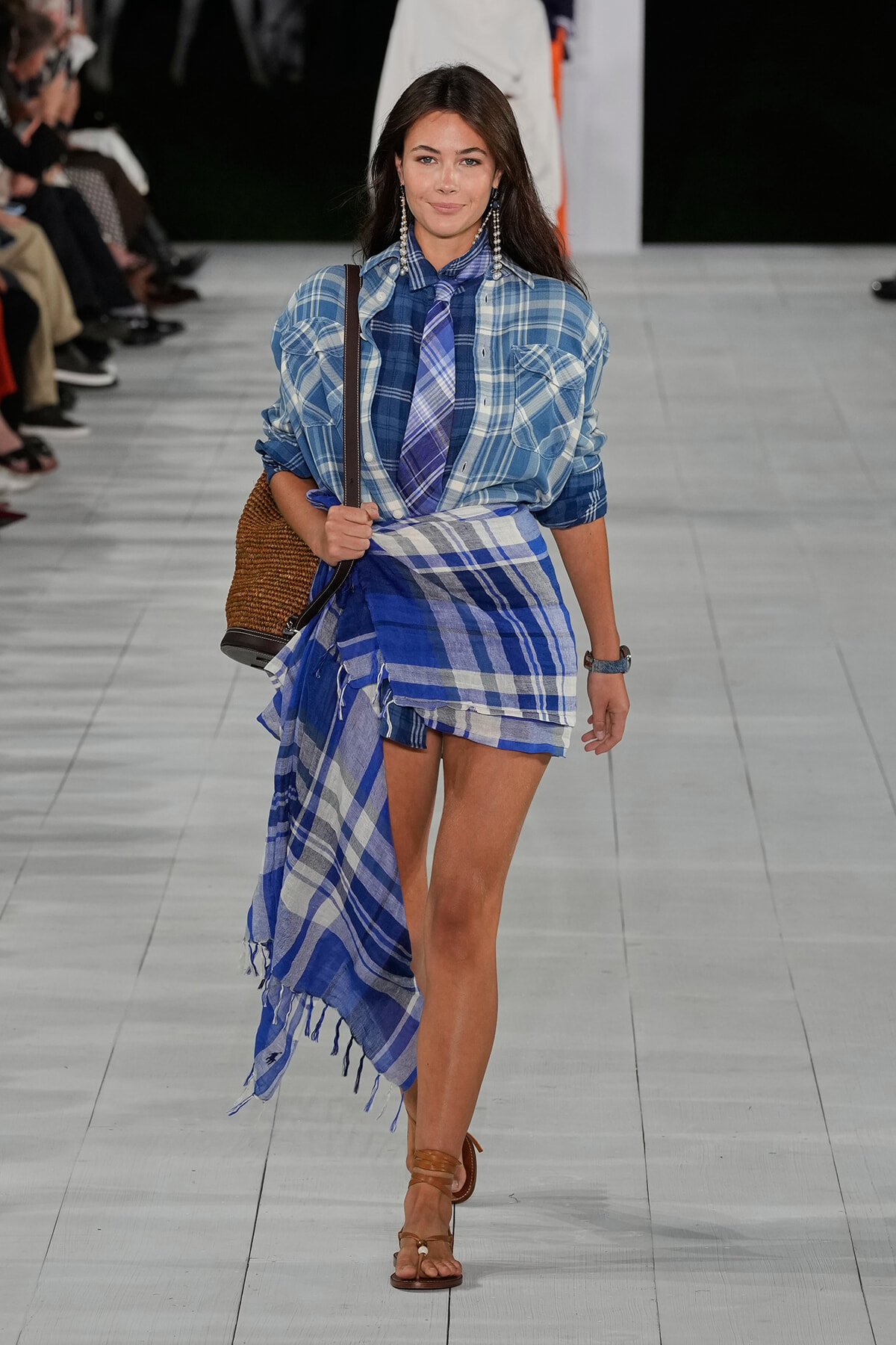 Model walking runway in blue plaid shirt paired with a matching blue plaid wrap skirt and tie, carrying a woven straw bag and wearing brown sandals.
