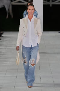 Model walking runway in a long cream blazer with white shirt and ripped light-wash jeans, holding a small cream handbag.