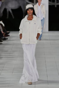 Model walking a fashion show in an all-white ensemble: white zip jacket, flowing white skirt, and a white cap.