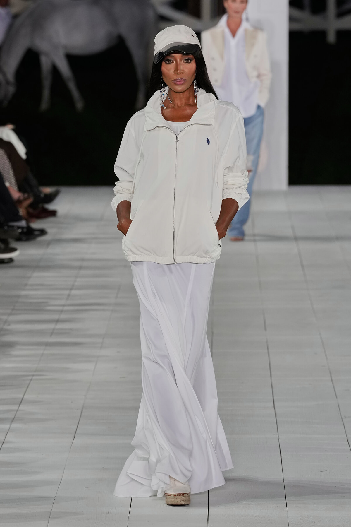Model walking a fashion show in an all-white ensemble: white zip jacket, flowing white skirt, and a white cap.