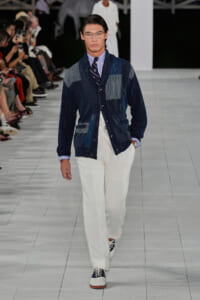 Male model walks the runway in a patchwork navy denim cardigan over a striped shirt and tie, paired with white trousers and two-tone sneakers; audience lines the catwalk.