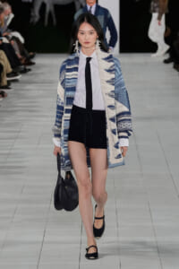 Runway model wearing a blue-white patterned oversized cardigan over a white shirt with a black tie, black shorts, and black sandals, carrying a black bag.