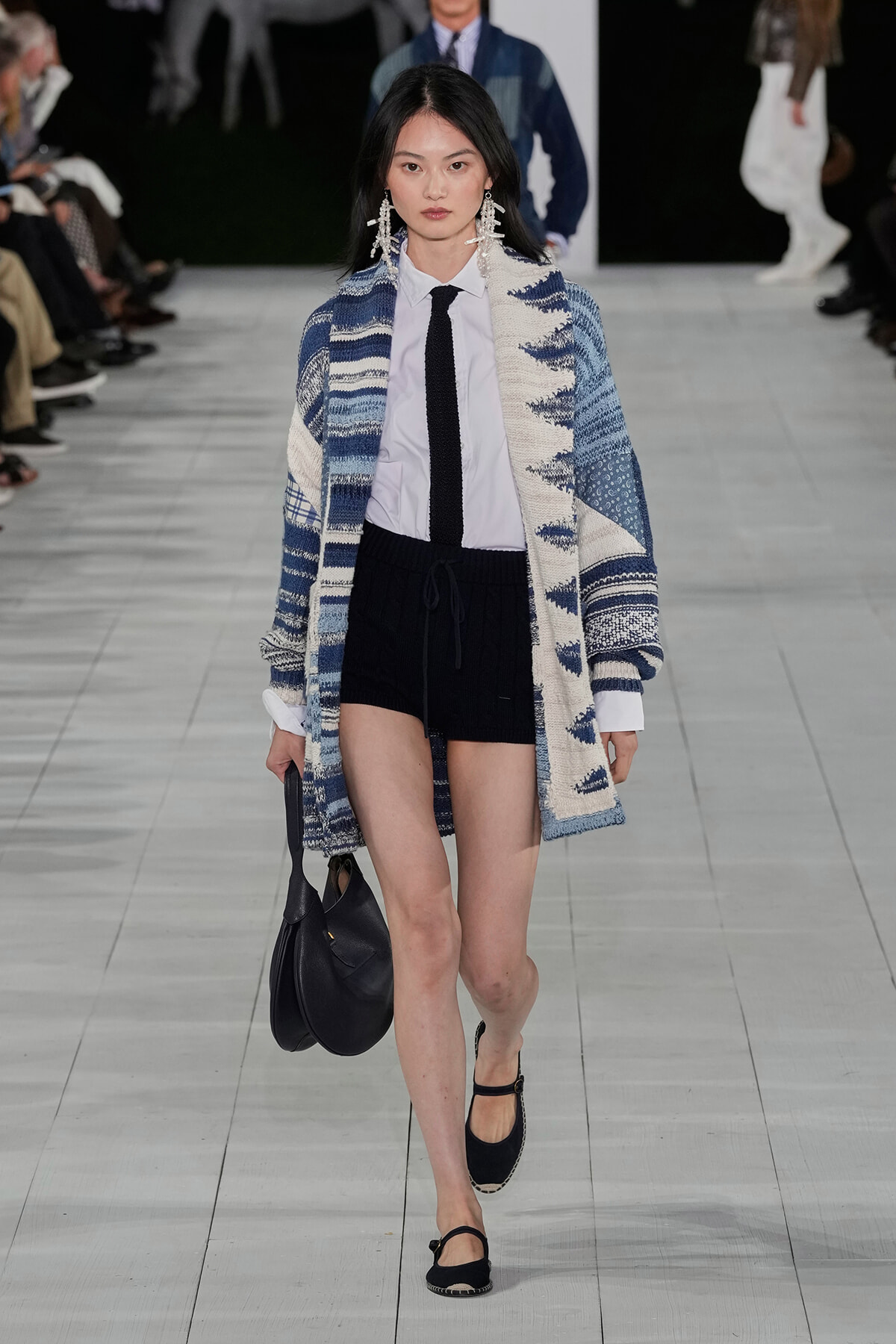 Runway model wearing a blue-white patterned oversized cardigan over a white shirt with a black tie, black shorts, and black sandals, carrying a black bag.