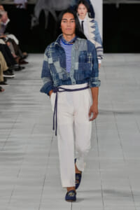 Model walking a runway in a patchwork denim shirt over a blue-and-white striped top, white wide-leg pants with a purple belt, and blue embroidered loafers.