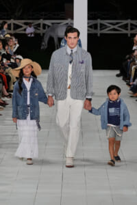 Model in a light striped jacket and white pants walks the runway, joined by a girl and a boy in denim jackets holding hands.