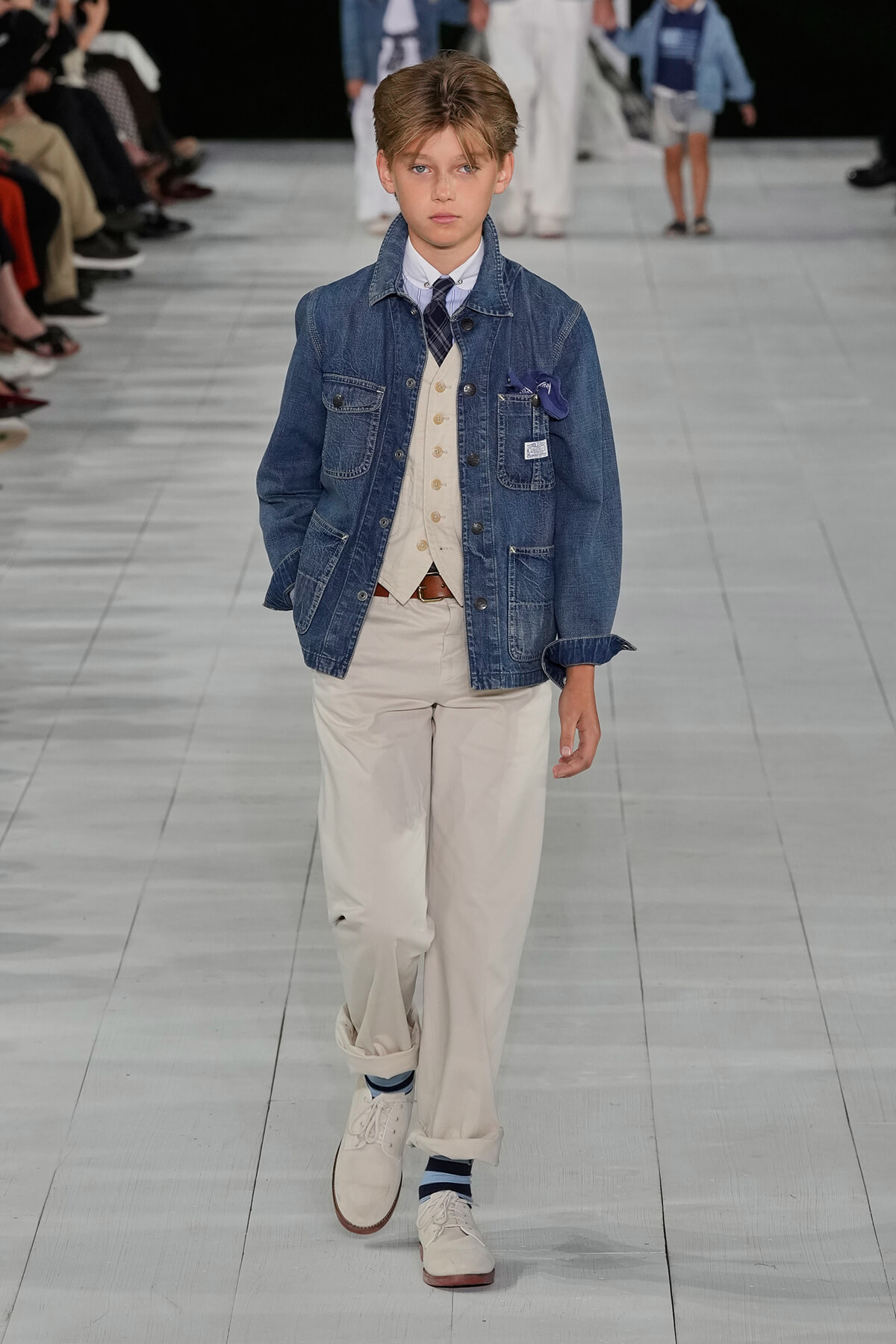 Young boy model walking a fashion runway in a denim jacket, beige vest, white shirt, and beige trousers with white shoes.