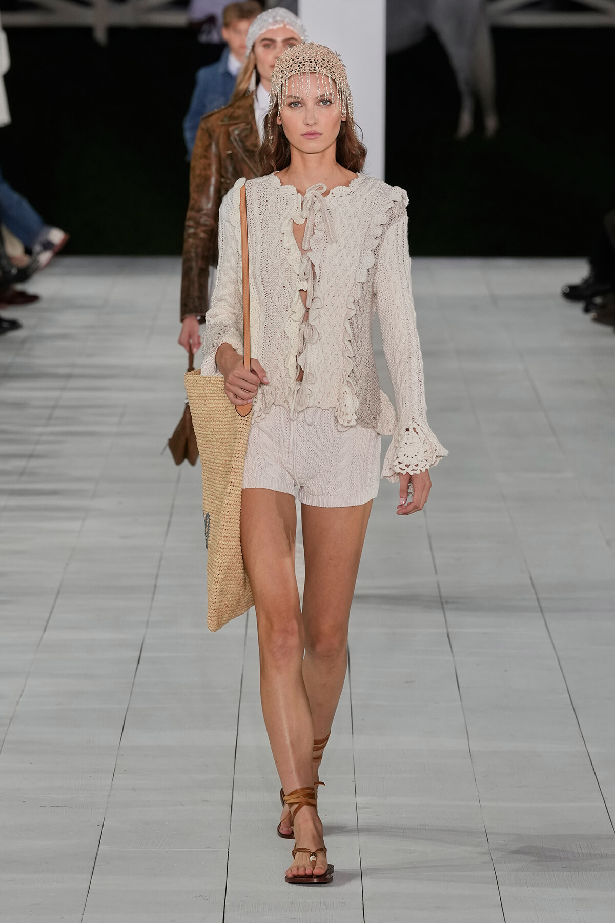 Model walking a fashion runway in a cream crochet blouse with tie-front and matching knit shorts, paired with a beaded headpiece and a large straw tote.