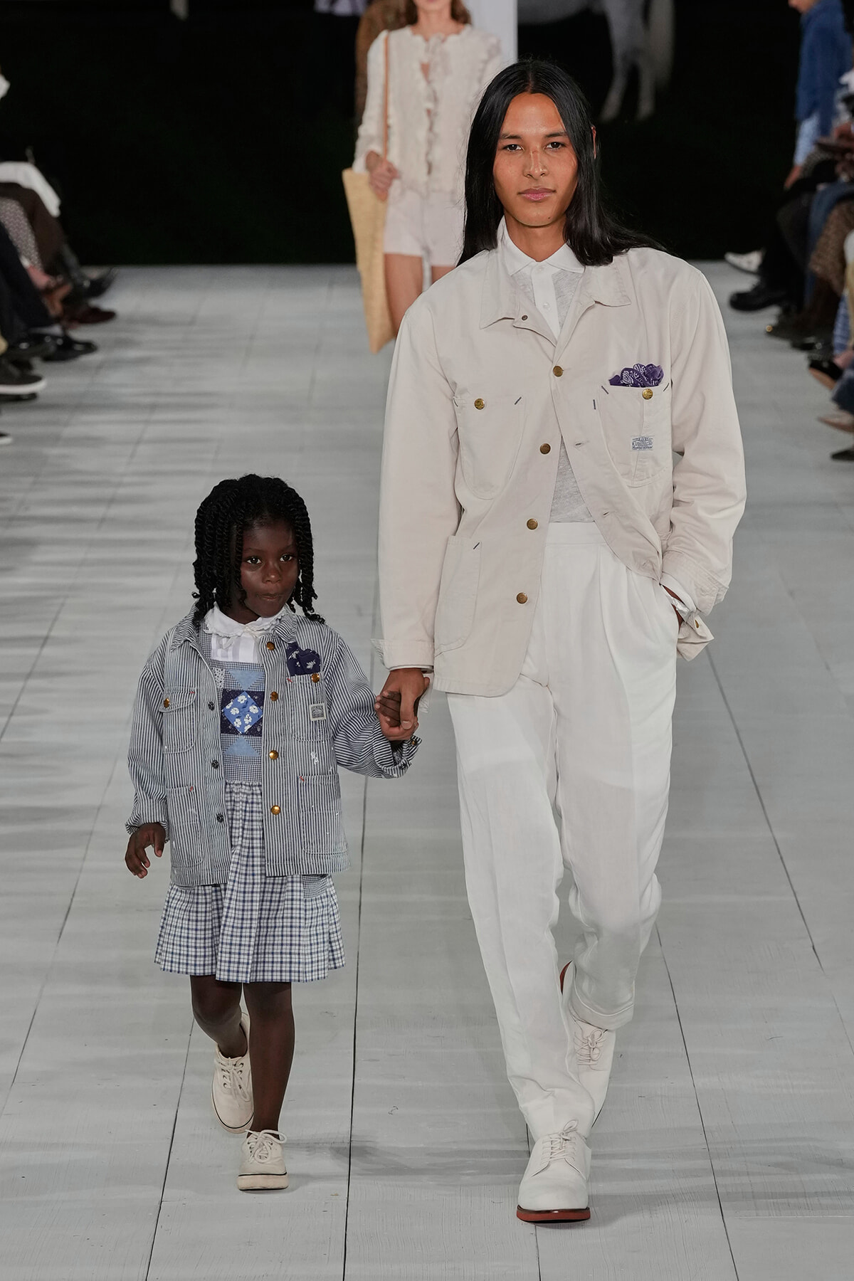 Adult and child model walk hand-in-hand down a fashion runway; both in light-colored outfits, the adult in a cream jacket and pants, the child in a striped jacket and checked dress.