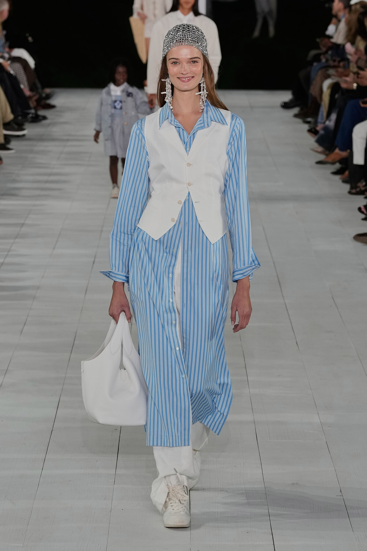 Model on a runway wearing a blue striped shirt dress with a white waistcoat, white boots, and a white tote, topped with a silver beaded cap.