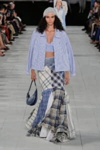 Model on the runway wearing a light blue striped jacket over a blue-striped bralette, paired with a patchwork plaid maxi skirt and a silver beanie, carrying a small blue bag.