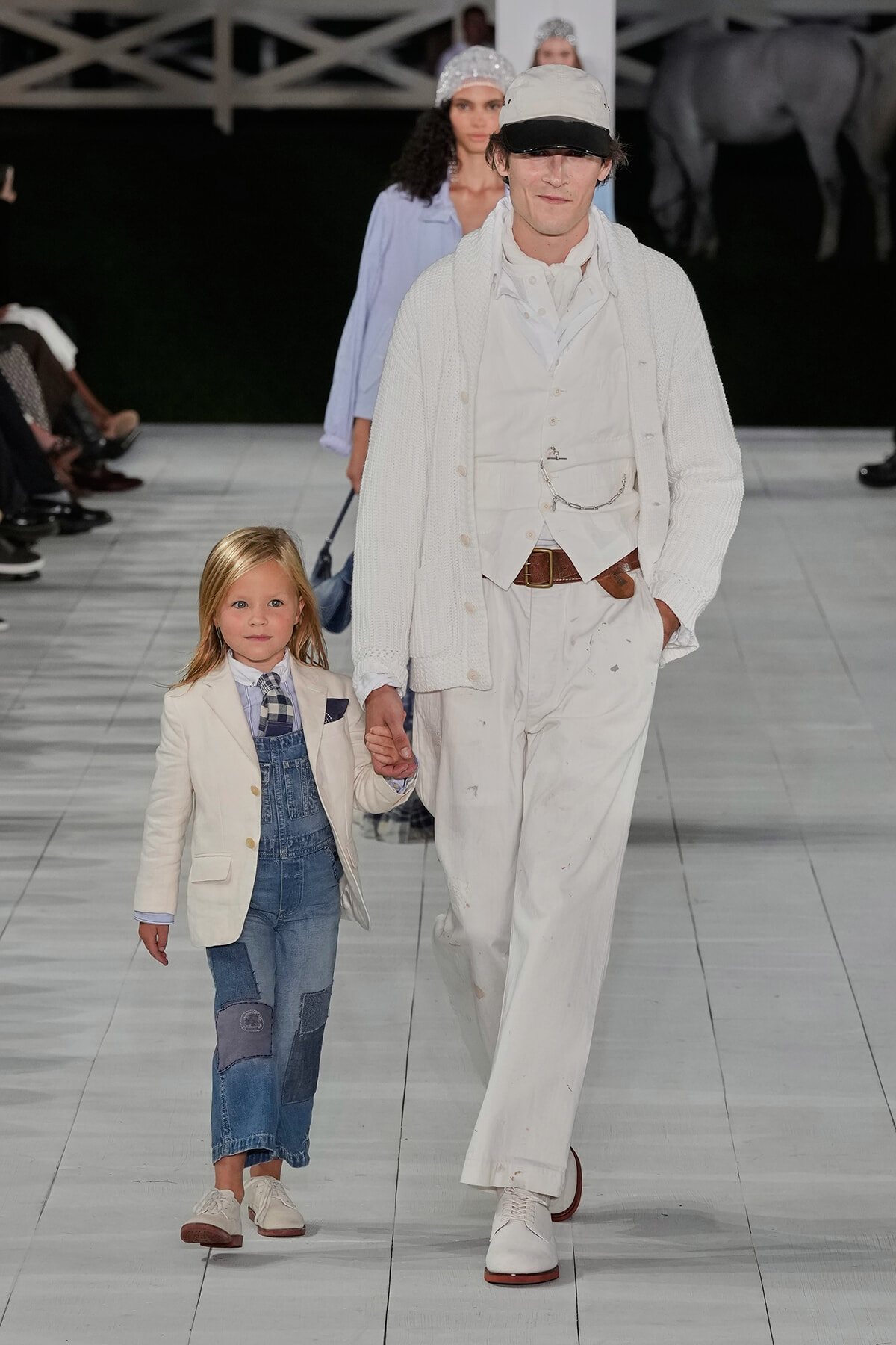 Male model in an all-white ensemble (knit cardigan, vest, cap) walks hand-in-hand with a young girl in a cream blazer and patchwork denim overalls on a fashion runway.