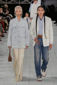 Two models walk a fashion runway side by side: one in a light denim jacket over a cream satin skirt, carrying a brown bag; the other in a white blazer, striped shirt, and jeans with braids and turquoise rings. Audience watches.