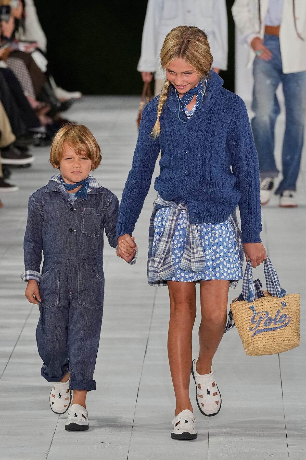 Two young children walk hand-in-hand down a fashion runway in blue outfits; girl carries a straw tote with 'Polo' on it.