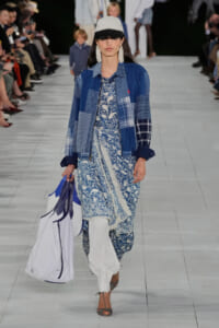 Female model walks runway in a blue patchwork jacket over a blue-and-white printed dress, white pants, gray cap, statement earrings, and a large white-and-blue tote bag.