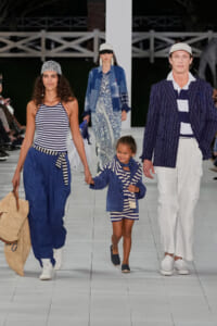 Runway scene with a woman in striped top and blue pants, a smiling girl in navy and white stripes, and a man in a pinstripe blazer walking the catwalk in nautical outfits.