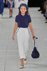 Young girl model walks a fashion runway in a navy knit polo and white high-waisted pants, carrying a navy drawstring bag.
