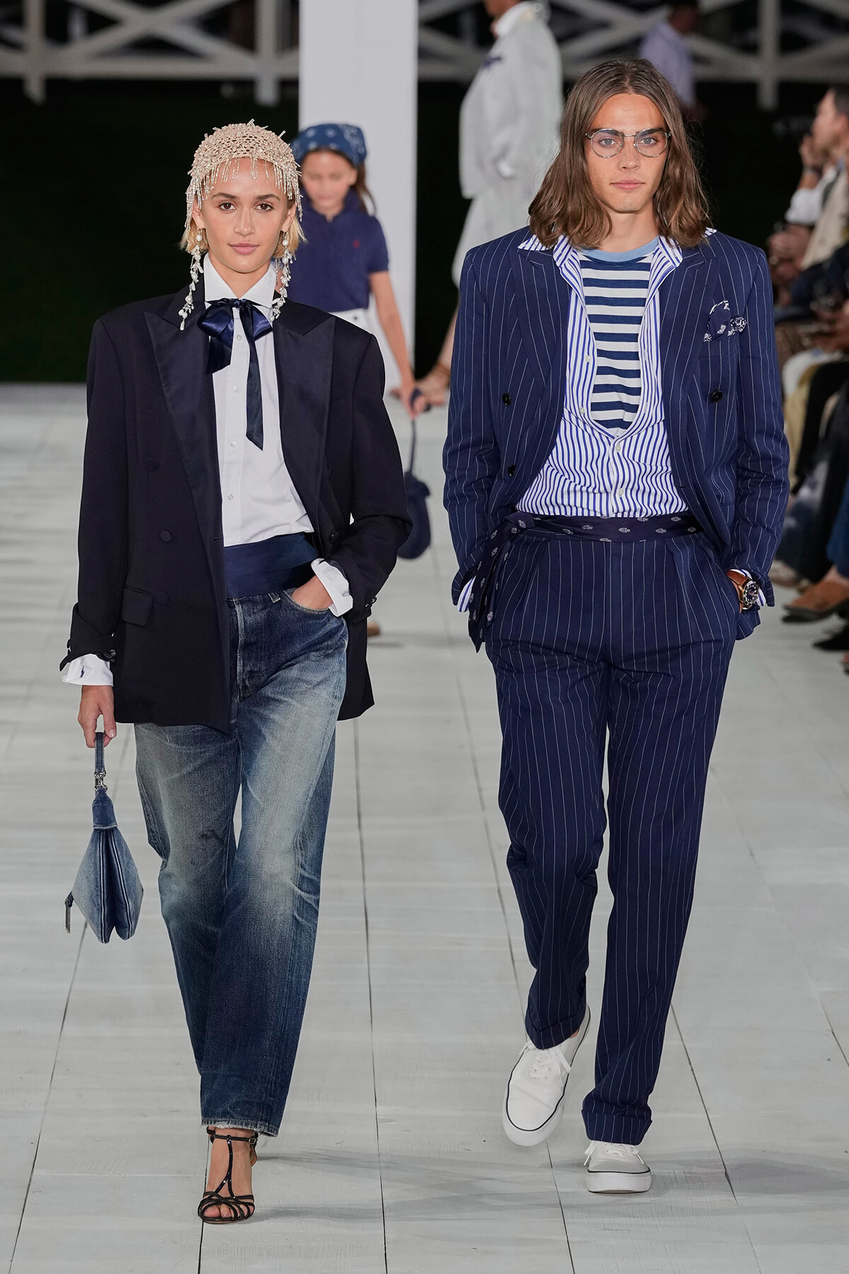 Two models walk a fashion show runway: left, woman in navy blazer, white shirt with bow, jeans, pearl headpiece, carrying a small blue bag; right, man in blue pinstripe suit with striped shirt and white sneakers.