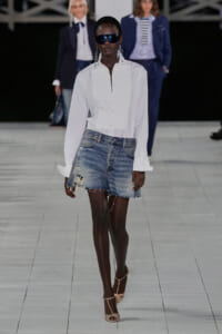 Model walking a fashion runway in a white button-down shirt tucked into distressed denim shorts, wearing sunglasses and strappy heels.