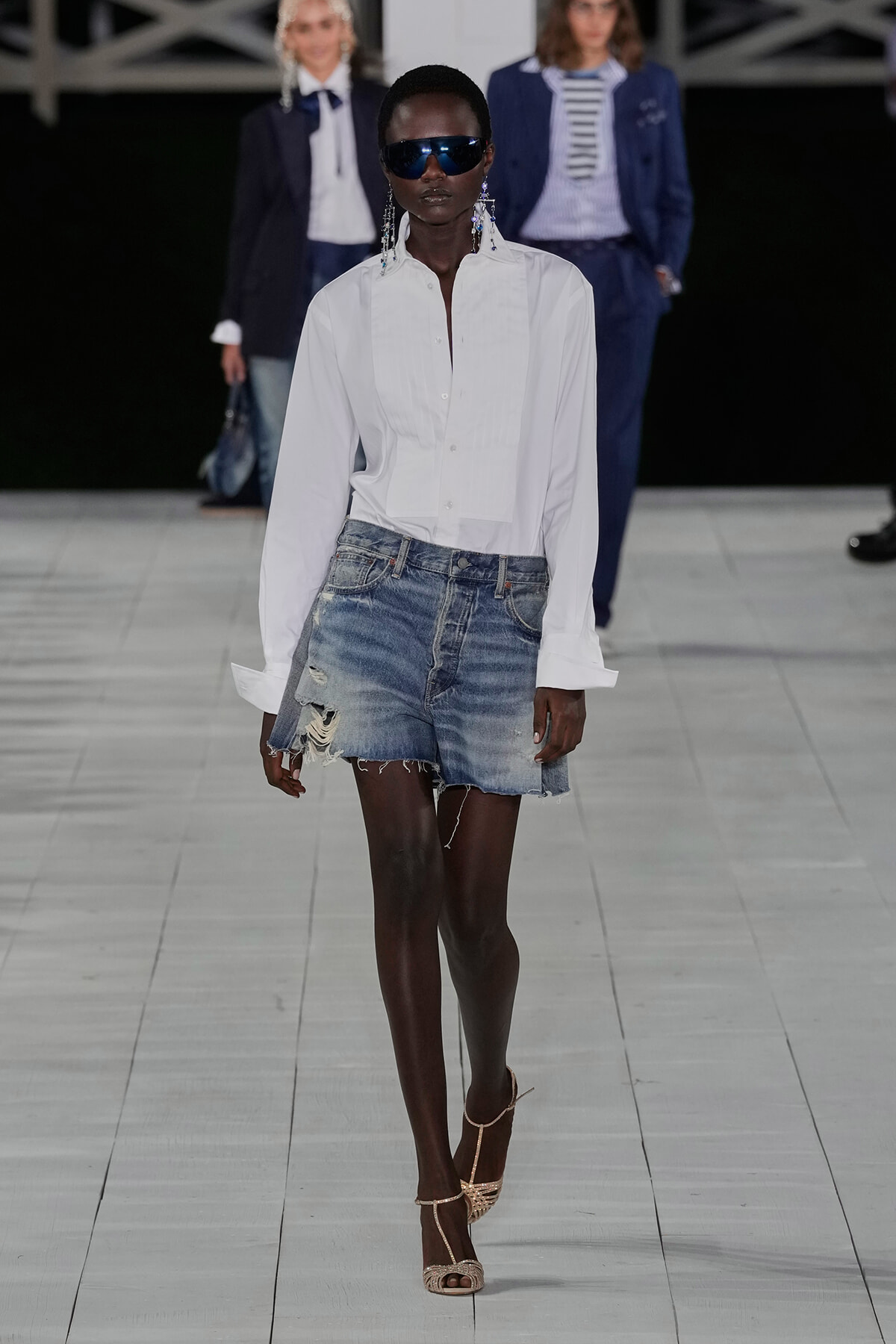 Model walking a fashion runway in a white button-down shirt tucked into distressed denim shorts, wearing sunglasses and strappy heels.