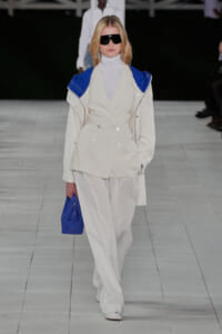 Runway model in a cream double-breasted suit with blue shoulder accents, white turtleneck, oversized black sunglasses, and a cobalt blue bag.