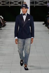 Male model on a runway wearing a navy double-breasted blazer, white shirt, black bow tie, white pocket square, distressed jeans, and navy NY cap with suede loafers showing NY logo