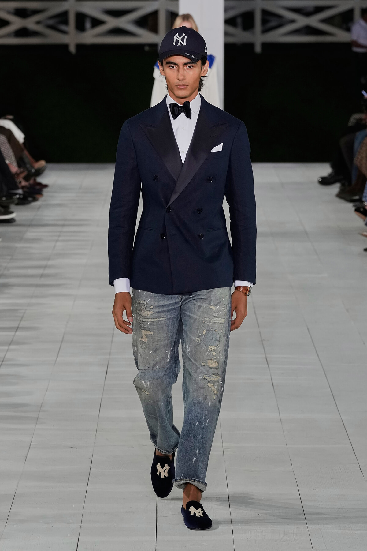 Male model on a runway wearing a navy double-breasted blazer, white shirt, black bow tie, white pocket square, distressed jeans, and navy NY cap with suede loafers showing NY logo